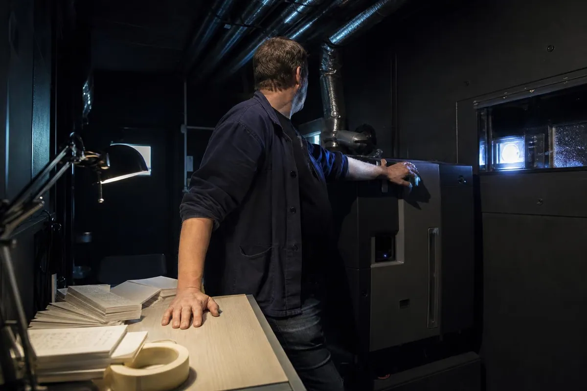 Un projectionniste vérifie le lancement de la séance dans une cabine de projection, la main sur le flanc du projecteur numérique